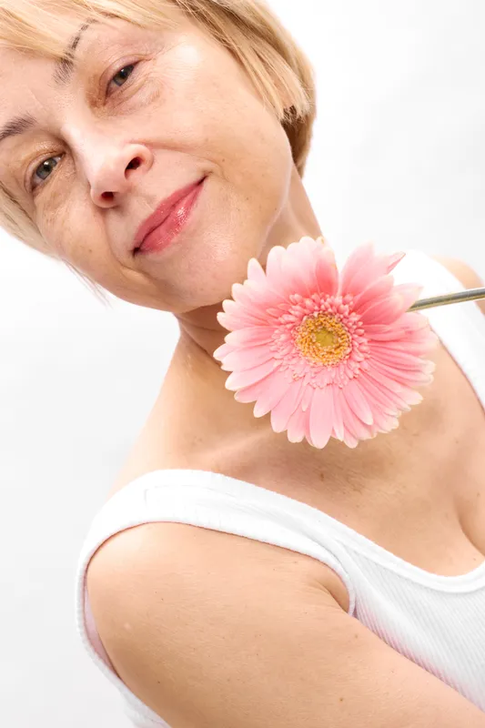 Mature woman portrait closeup with pink daisy flower near lips, soft makeup highlights beauty and freshness, calm spa mood, white background, studio smiling elegance