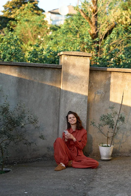 Casual outdoor lifestyle shot featuring a woman in red sitting on the ground by a courtyard wall, enjoying a drink with a warm relaxed mood and natural sunlight