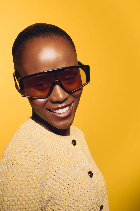 Smiling young black woman with dark skin wearing large stylish sunglasses and a yellow knit sweater on yellow background. Concept of fashion, summer, and joyful mood in portrait photography.