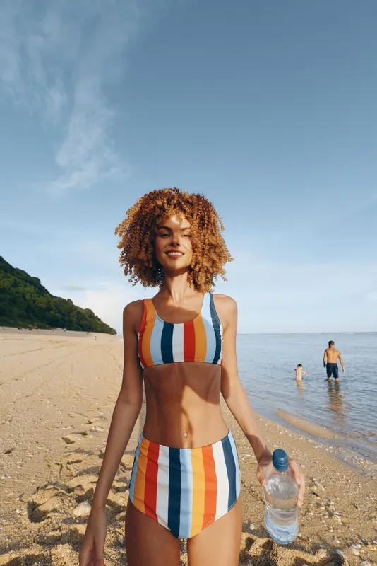 Young woman with curly hair wearing striped swimsuit standing on sandy beach holding water bottle smiling under clear blue sky sunny day relaxing outdoors