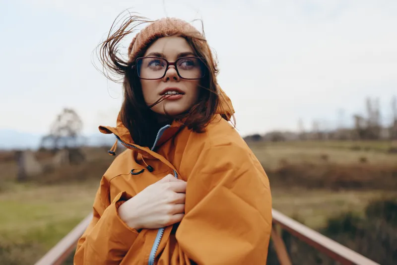Woman lifestyle outdoors portrait in autumn, orange jacket on a countryside road, wind lifting her hair, thoughtful gaze, soft daylight, warm colors, calm travel mood
