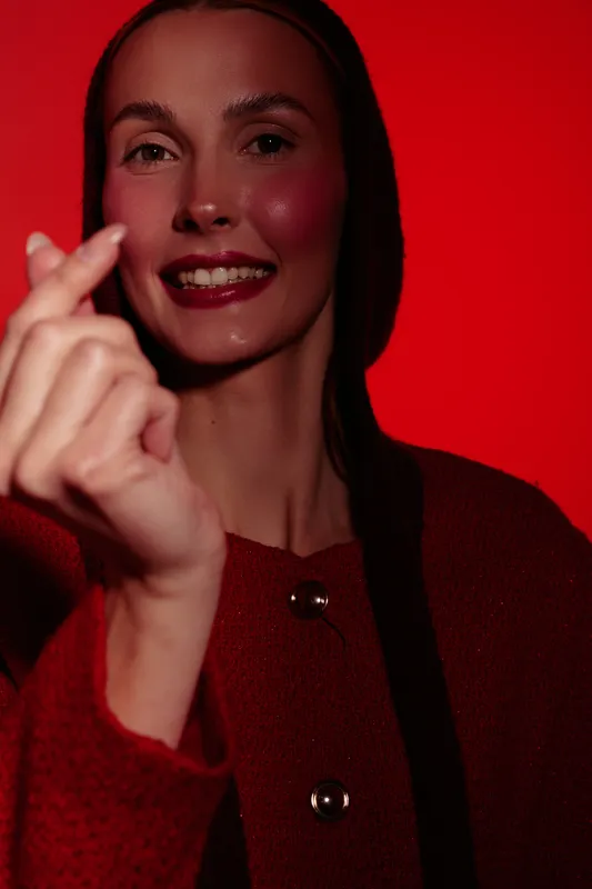 Smiling woman with long dark hair makes finger heart gesture against red background. Close-up portrait with warm lighting, conveying happiness, affection, and positive emotions in studio setting.