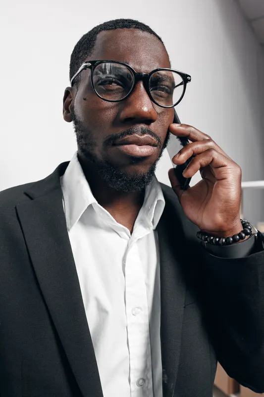 African American man with dark skin wearing glasses and black suit talking on smartphone in modern office interior with natural light and white walls.