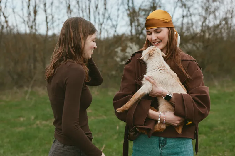 Goat kid woman outdoors scene shows friendship, nature, and joy as two people smile while a playful animal is held in warm light, capturing lifestyle and companionship