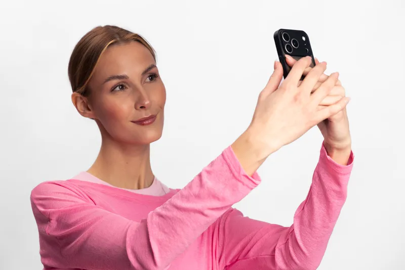 Young woman in pink sweater takes selfie with smartphone. She smiles slightly in bright studio on white background. Concept of social media, communication, and modern lifestyle.