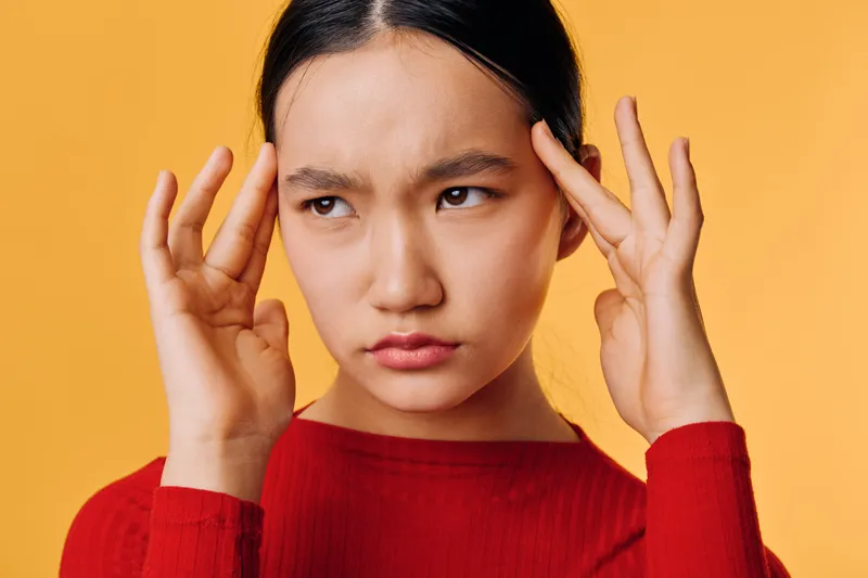 Young woman with dark hair in red sweater touching temples with fingers, showing worried or stressed expression. Portrait on orange background with focus on face and emotion.