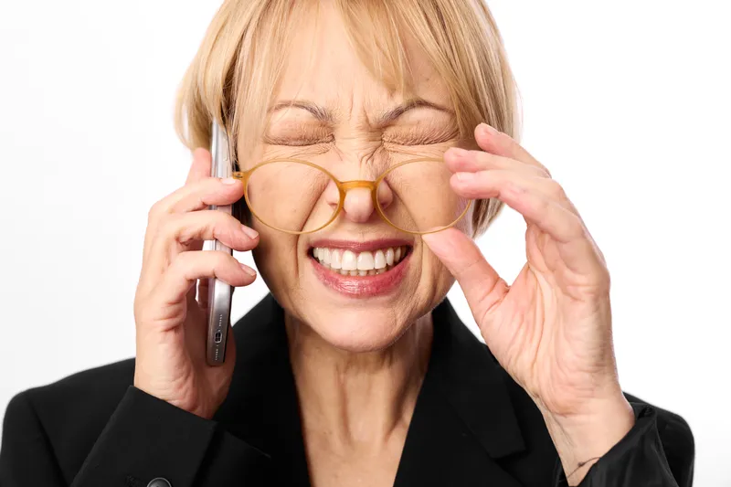 Smiling woman elderly adjusts glasses while holding phone to her ear, business portrait on white background, studio light highlights face, communication concept.