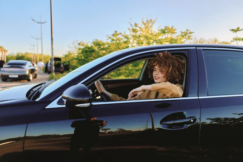 A stylish woman driver in a dark car enjoys a sunlit ride with retro film color tones, capturing a candid lifestyle moment on the road.