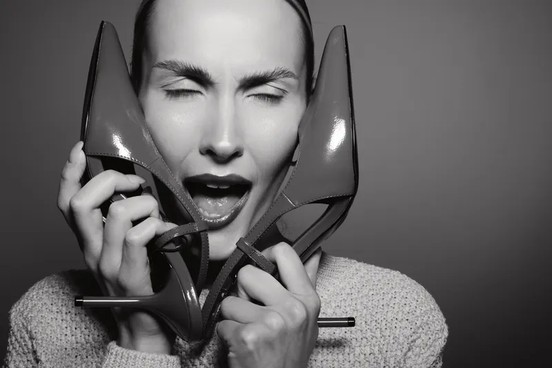 Young white woman with pale skin holds high heel shoes near her face with closed eyes and open mouth, expressing emotion. Studio portrait in black and white.