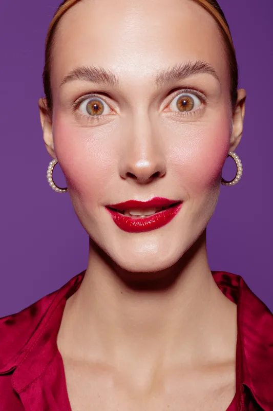 Close-up portrait of surprised woman with wide eyes and red lipstick wearing hoop earrings and red blouse on purple background. Concept of emotion, beauty, and fashion expression.
