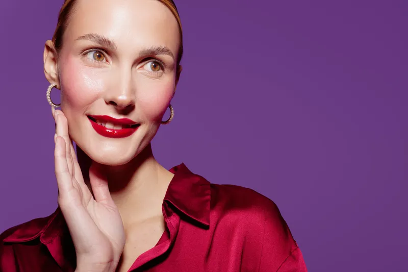 Portrait of a smiling woman with red lipstick and shiny skin wearing a red satin blouse and hoop earrings on purple background. Concept of beauty, fashion, and elegance.