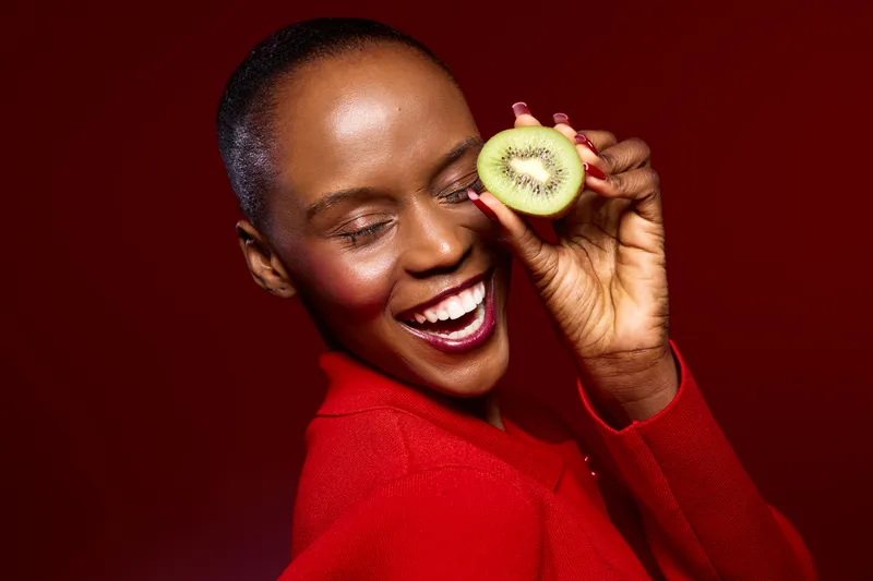 Happy African woman with dark skin wearing red sweater holds half of a kiwi fruit near her eye smiling with closed eyes. Concept of healthy lifestyle, fruit, and joyful emotions.