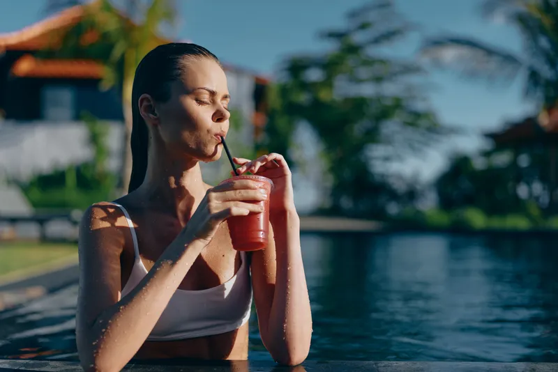 A young woman in a white swimsuit sips a pink drink through a straw while lounging at a sunlit pool, conveying a carefree, summery moment.