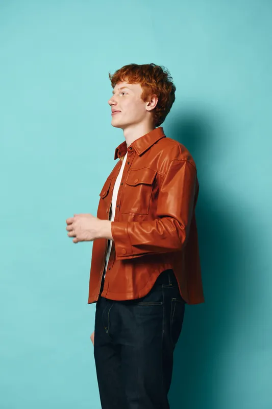 Young man with curly red hair wearing brown leather jacket and dark jeans standing sideways against blue background. Casual outfit, fashion style, portrait and confident expression.