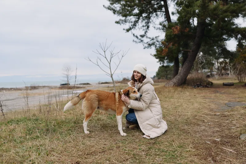 Woman kneels beside a lake with a fluffy dog in chilly weather, wearing a long coat and knitted hat, trees and shore in the background