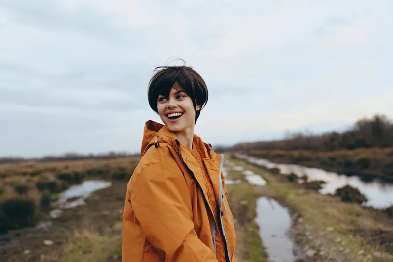 Woman lifestyle outdoors, smiling on hiking path, wearing orange jacket, standing near narrow trail and river, cloudy sky, scenic nature background, autumn mood, travel portrait