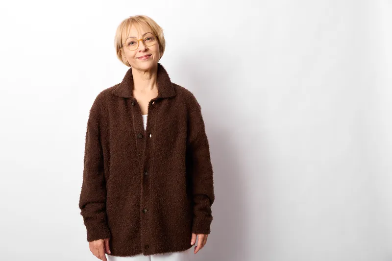 Senior woman portrait, smile and calm expression, studio white background, brown jacket, glasses, relaxed posture, casual fashion, indoor scene, copy space
