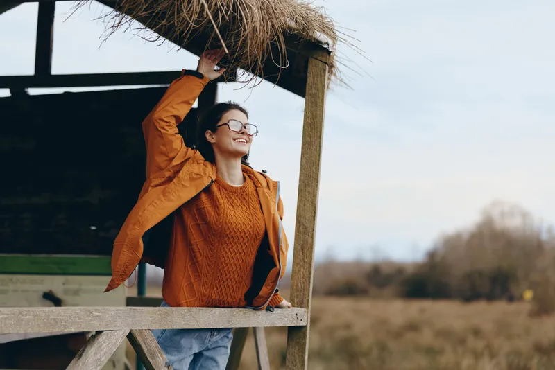Woman lifestyle outdoor portrait, smiling with glasses at wooden platform in spring countryside, fresh air, warm sunlight and sky, sweater, calm wellness mood, natural background