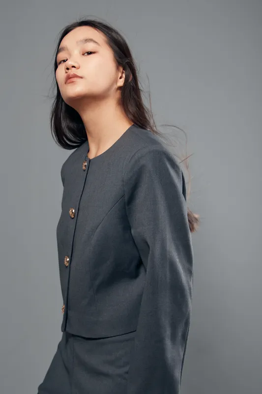 Young woman wearing dark blazer posing with confident expression in studio with plain gray background, long hair loose and natural, professional style portrait of female model.