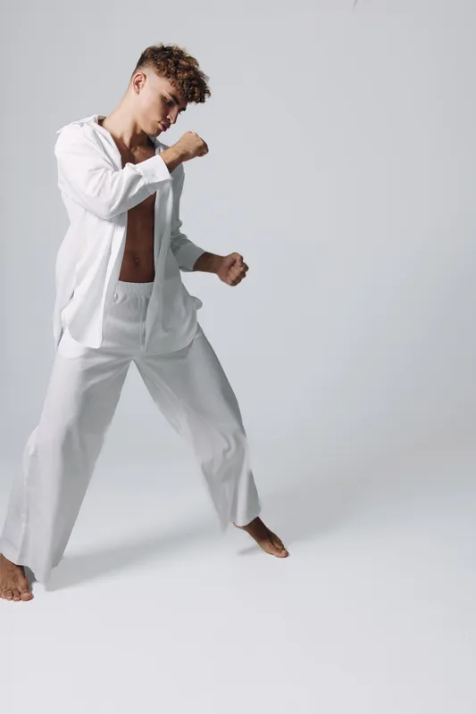 Young man in white loose clothing practicing martial arts stance barefoot on plain background with focused expression and curly hair in studio setting.