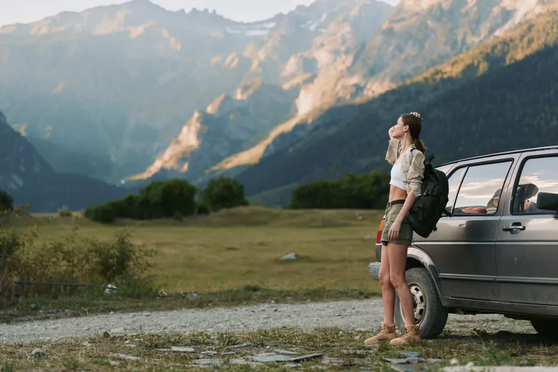 Woman traveler beside a car in a mountainous landscape, backpack ready, gazing at distant peaks, outdoor adventure, nature scenery, bright daylight, calm mood.