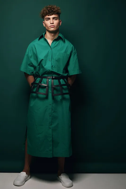 Young man standing against dark green background wearing oversized green shirt dress with black belt and white sneakers, posing with hands behind back in studio lighting.