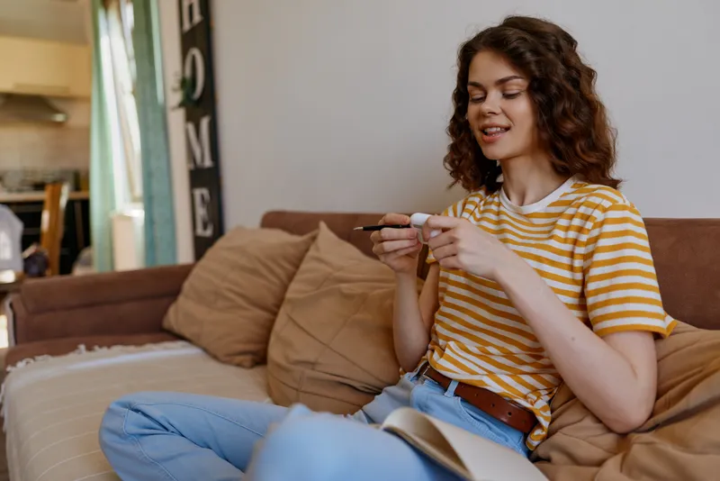 Woman sits on a cozy sofa at home, casually using a smartphone, wearing a striped shirt and jeans, relaxed mood, bright natural light, everyday indoor lifestyle, with a calm, cozy vibe.