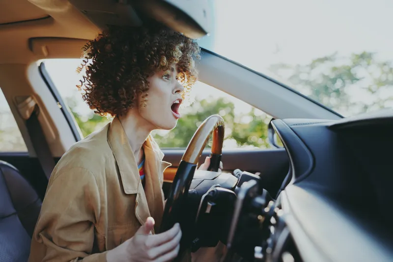 Young woman with curly hair drives a car, shouting with excitement as sunlit light streams through the windshield, capturing a candid road moment.