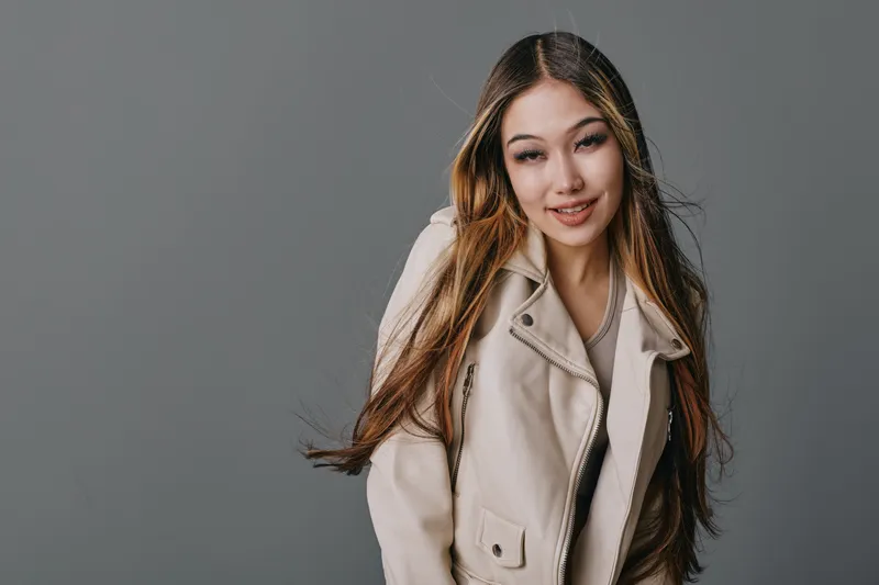 Young woman with long hair smiling and wearing beige jacket posing on gray background. Female model with natural makeup and casual outfit for fashion or lifestyle concept.