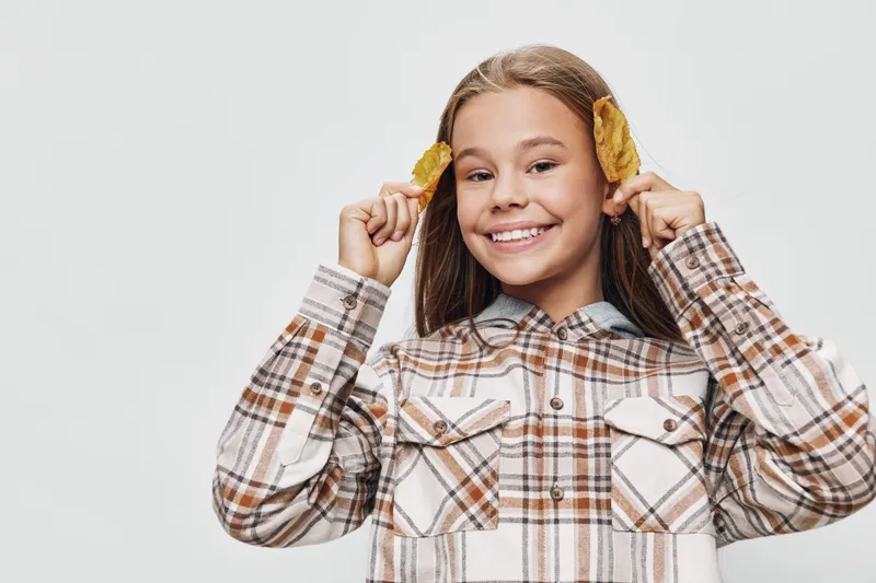 Smiling girl with long hair wearing checkered shirt holding dried flowers near ears. Happy child posing on white background with playful expression and joyful mood in studio portrait.