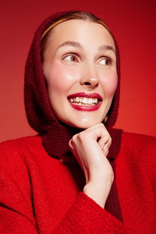 Smiling woman in red sweater and headscarf looks away with bright lipstick on red background. Concept of fashion, beauty, happiness, and winter style portrait with warm colors.