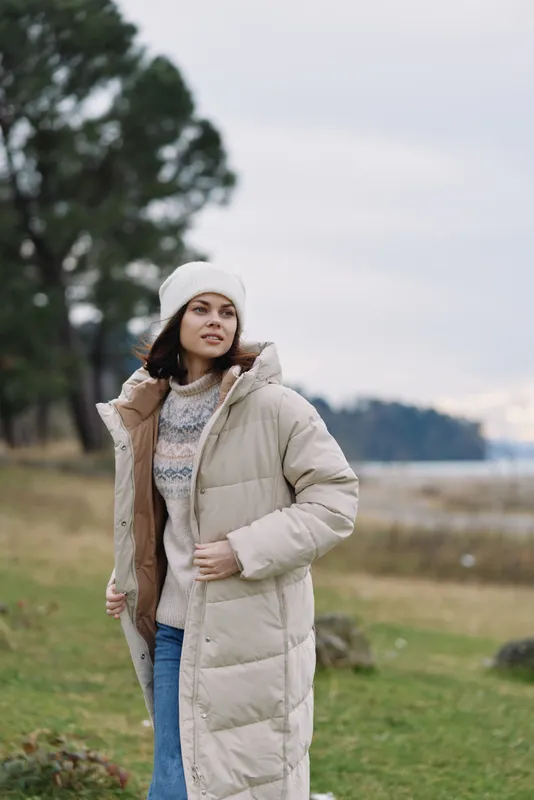 Woman outdoors in a winter coat and puffer jacket in a quiet landscape, wearing a knit sweater and beanie, soft natural light and calm mood for a seasonal fashion moment.