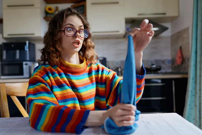 A woman in a colorful striped sweater irons a blue cloth at a kitchen table, bright home scene highlighting everyday casual chores and relaxed domestic life, with soft daylight in a warm interior.