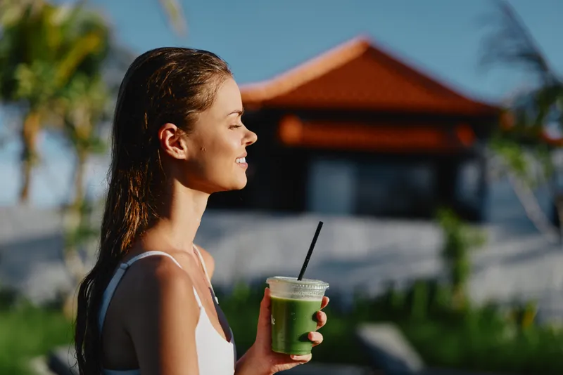 Young woman outdoors holding green smoothie cup, enjoying sunny day, casual outfit, relaxed expression, natural light, tropical background with palm trees and house.