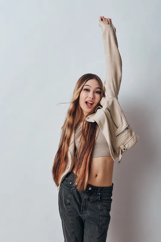Young woman with long hair wearing casual jacket and crop top posing happily with raised arm against white background in studio. Fashion, beauty, and lifestyle concept.