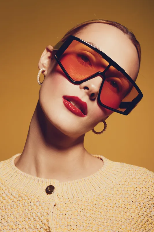 Fashionable woman with oversized orange sunglasses and red lipstick poses against yellow background. She wears a textured beige sweater and hoop earrings. Concept of modern style and beauty.
