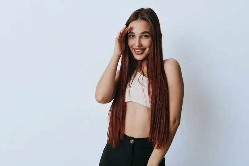 Young woman with long braided hair smiling and posing with hand near face against white background. Casual outfit with crop top and black pants in studio portrait.