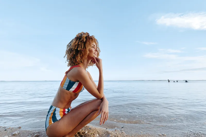 Woman on beach, sitting in colorful striped swimsuit, profile pose, touching cheek, calm ocean horizon, sunlight summer sky, gentle breeze, natural light, relaxed serene seaside tranquil