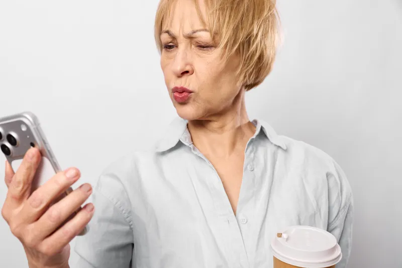 Woman holding smartphone, showing anger and stress, close up facial expression with short blond hair, holding a takeaway cup, bright studio background, modern mobile communication