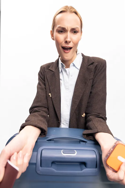 Surprised young woman in brown blazer holds blue suitcase and passport, looking down with astonished expression. Concept for travel, packing, and unexpected situations.