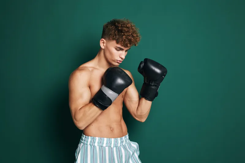 Young man with curly hair wearing boxing gloves and striped shorts training in front of green background for fitness and martial arts practice.