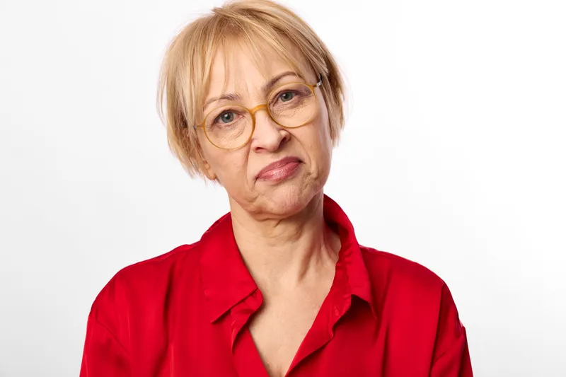 Senior woman portrait in studio, wearing red blouse and round glasses, short blond hair, calm face, looking at camera against bright white background