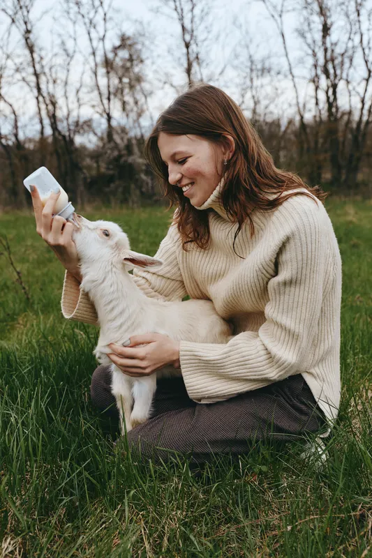 Caring woman seated on grass bottles feeds a young goat kid, tender moment outdoors, rural setting, cozy sweater, gentle interaction, natural light, warm mood, wholesome family style scene during