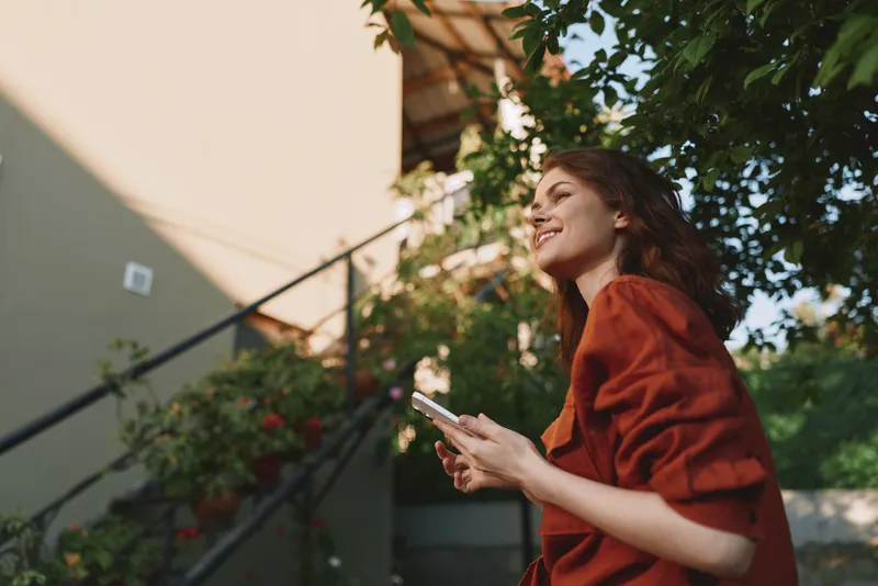 Woman lifestyle shot outdoors, smiling woman in a red blouse holds a smartphone, enjoying a sunny day in a garden with stairs and greenery, candid everyday moment, bright natural light
