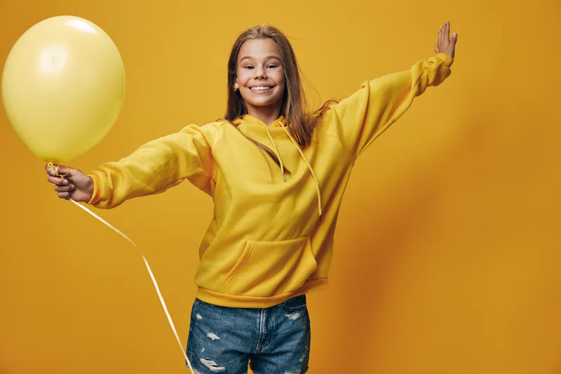 Girl with long hair wearing yellow hoodie holding balloon and smiling standing with arms outstretched against yellow background for celebration and joy.