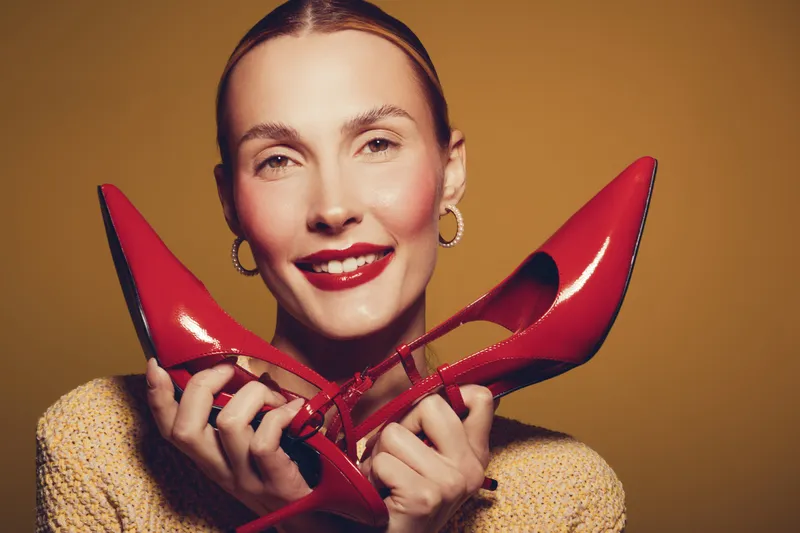 Smiling woman with red lipstick holds red high heels near face against brown background. Concept of fashion, style, beauty, and elegance with bright studio lighting and joyful mood.