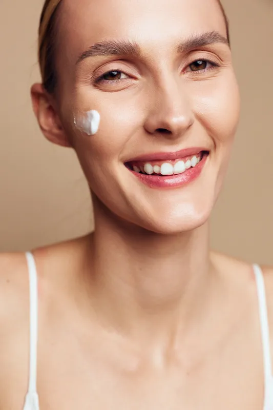 Smiling young woman with a dab of white cream on her cheek poses for a close-up portrait, highlighting skincare, self-care, and healthy skin in a soft beige studio setting.