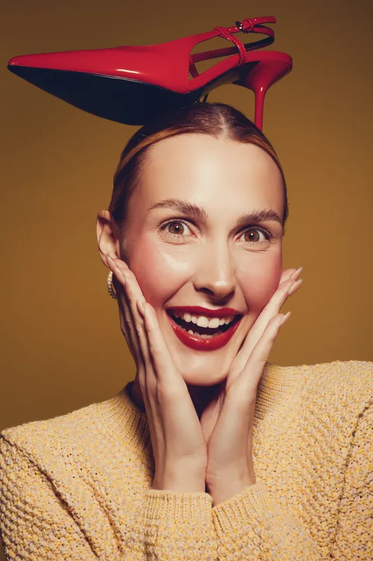 Smiling woman with red high heel shoe on head expresses surprise and joy. She wears a yellow sweater and bright lipstick, isolated on warm brown background. Concept of fun and fashion.