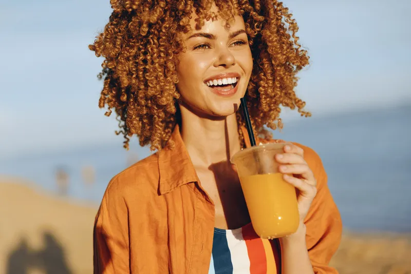 Curly hair woman smiles while holding a beverage with straw, orange drink in hand, seaside summer coast background, warm sunlight, casual jacket, shallow depth portrait natural outdoors