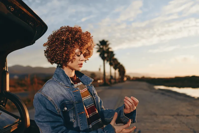Woman lifestyle outdoors sunset, curly hair denim jacket scarf by car, evening road walk, relaxed posture, warm golden light, coastal scenery, natural colors, soft breeze